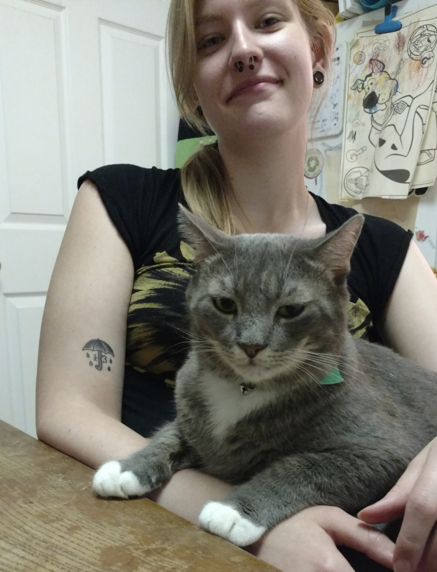 Savanna sitting with a grey cat in her lap at a brown wooden table in front of a fridge with colored illustrations on it.