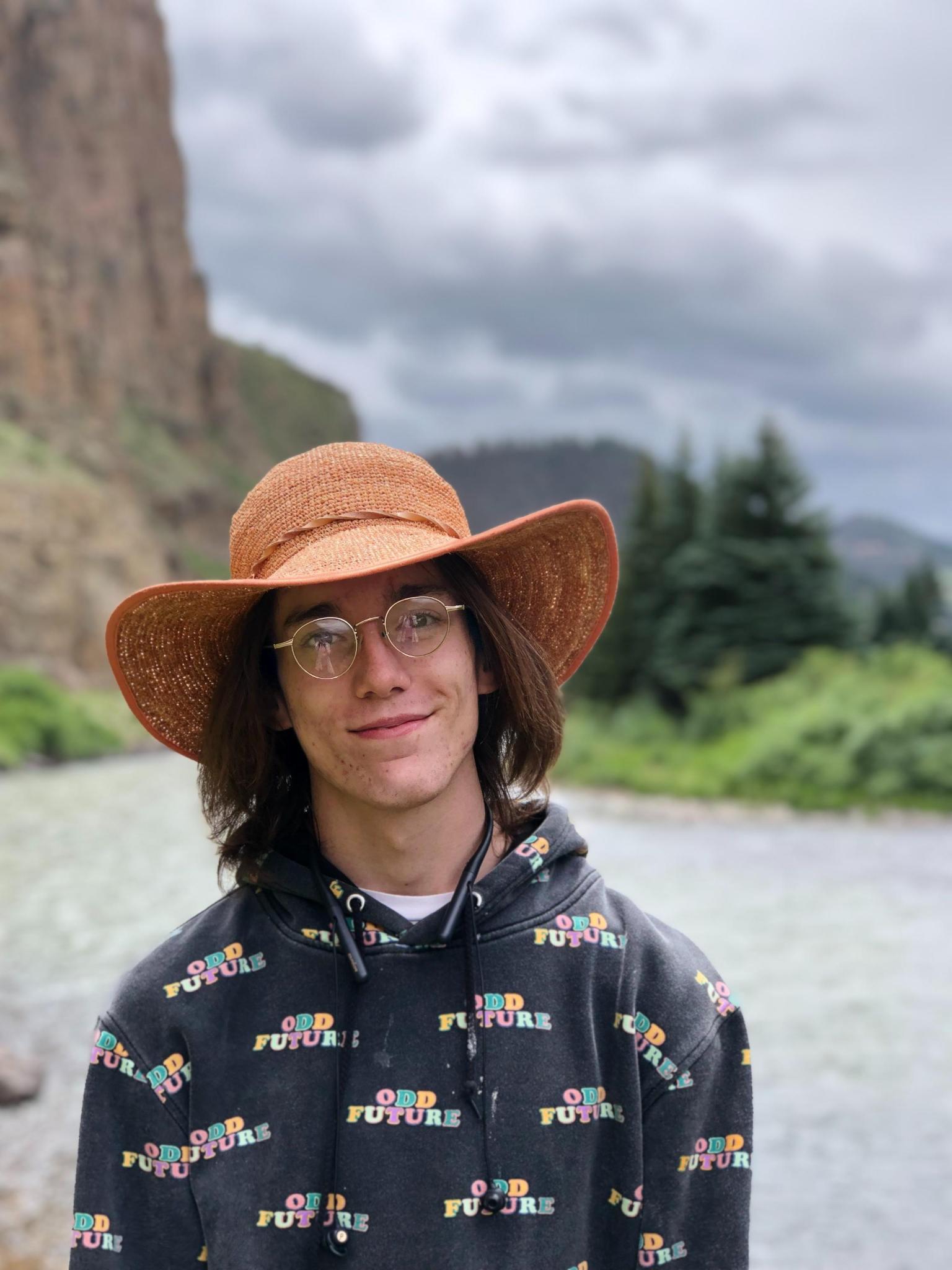A white woman with dark hair and glasses, wearing a wide-brimmed hat smiling while on a hike in a canyon.